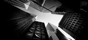 Black and white upward view between modern city skyscrapers toward a bright open sky
