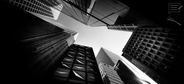 Black and white upward view between modern city skyscrapers toward a bright open sky