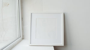 Blank white square frame held beside a window, representing the balance and simplicity of square artwork prints