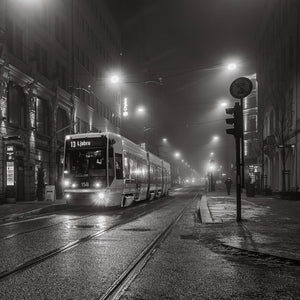 A tram on wet tracks at night, with overhead wires and streetlights fading into fog, in black and white.