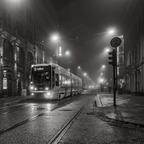 A tram on wet tracks at night, with overhead wires and streetlights fading into fog, in black and white.