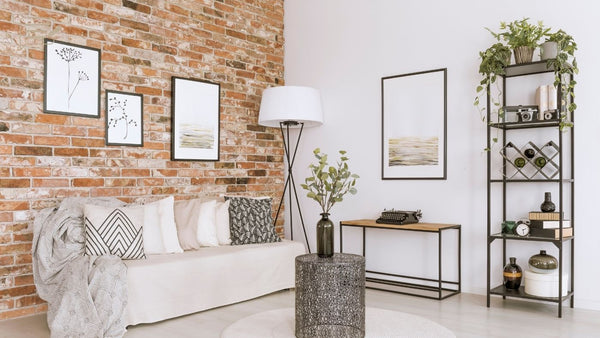 Living room with a white sofa, exposed brick, framed botanical and landscape artwork, a floor lamp and open shelving with plants.