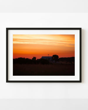 American farmhouse silhouetted beneath an orange and gold dawn sky.