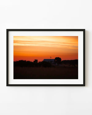 American farmhouse silhouetted beneath an orange and gold dawn sky.
