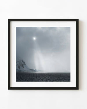 Veiled sun and shafts of light above a snowy Antarctic coast and dark sea.