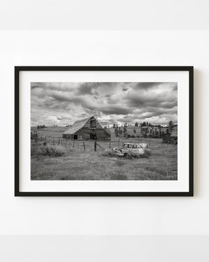 Abandoned Alberta barn and old car in tall grass beneath heavy clouds, in black and white.