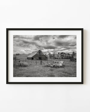 Abandoned Alberta barn and old car in tall grass beneath heavy clouds, in black and white.