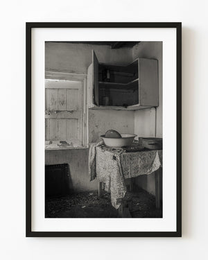 Abandoned kitchen in Clarence Town, Bahamas, with open cupboards and a cloth-covered table, in black and white.