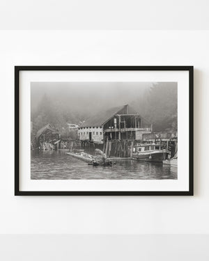 Abandoned British Columbia boathouse and moored boat beneath a misty forest, in black and white.