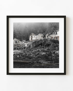 Abandoned wooden buildings above a rough British Columbia shore beneath a misty forest.