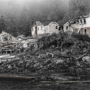 Square black and white photograph of weathered coastal houses and fallen timber in British Columbia.