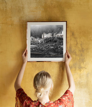 Pale abandoned buildings with pitched roofs above debris along a coastal shore.