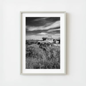 Framed black and white photograph of an abandoned car and structure in a field with a cloudy sky. 