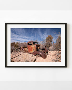 Rusted abandoned pickup truck in desert scrub beneath a streaked blue sky.