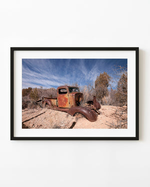 Rusted abandoned pickup truck in desert scrub beneath a streaked blue sky.