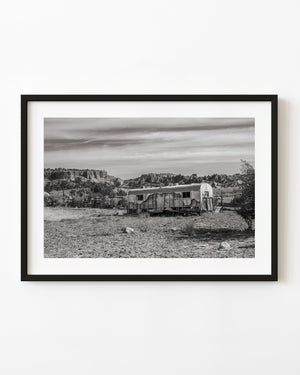Abandoned desert trailer beneath rocky hills, photographed in monochrome.