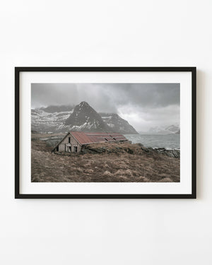 Abandoned Lofoten boathouse with a faded red roof beneath snowy mountains in Norway.