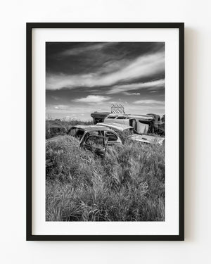Abandoned car bodies in tall Manitoba prairie grass beneath sweeping clouds, black and white