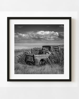 Abandoned Saskatchewan farm truck surrounded by tall grass beneath broad clouds, black and white