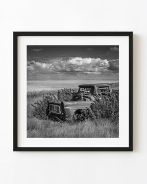 Abandoned Saskatchewan farm truck surrounded by tall grass beneath broad clouds, black and white