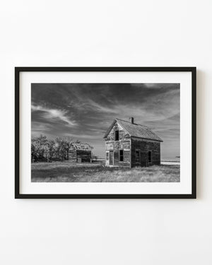Abandoned Saskatchewan farmhouse and smaller outbuilding beneath sweeping clouds, black and white