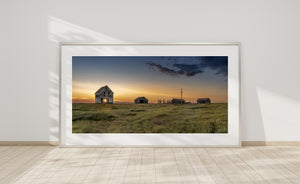 Weathered farm buildings stretch across grassy prairie beneath a gold and orange horizon.