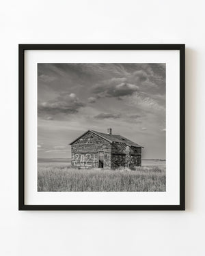 Weathered Saskatchewan shack beneath sweeping clouds in black and white.
