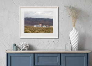 Low scrub surrounds an abandoned soda ash plant beneath a clouded mountain sky.
