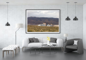 Desert vegetation leads toward pale factory buildings and distant California mountains.