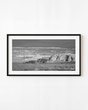 Abandoned Southwest farmstead and windmill beneath pale cliffs in black and white.