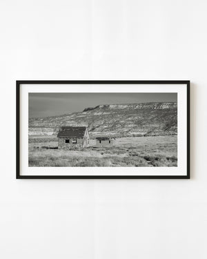 Two abandoned Southwest buildings beneath a long rocky ridge in black and white.