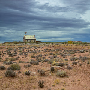 Square colour landscape photograph of an isolated schoolhouse in open desert.