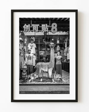 Alpaca sculpture among graphic shirts and reflections in a Beijing shop window, photographed in black and white.