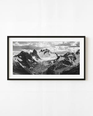 A pale alpine glacier lies between jagged dark mountain peaks in a black and white panorama.