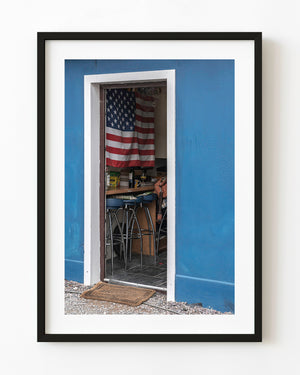 An American flag hangs inside a small back room seen through a white-trimmed doorway in a bright blue wall.