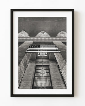 An American flag hangs above a chandelier and tall windows in Grand Central Terminal, photographed in black and white.