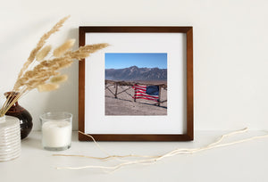 Narrow fence-post shadows cross dusty ground below a weathered American flag.
