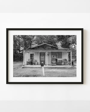 A weathered rural American house with porch chairs and a leaning mailbox is photographed in black and white.