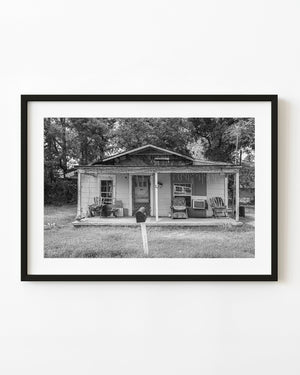 A weathered rural American house with porch chairs and a leaning mailbox is photographed in black and white.