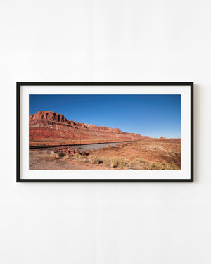 A railway follows layered red cliffs beneath a blue sky in a wide American Southwest colour photograph.