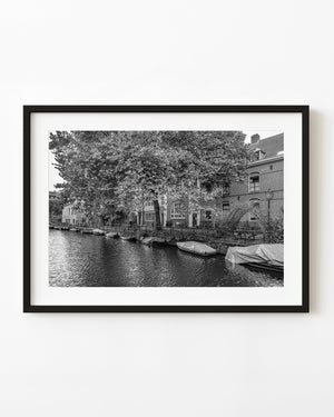 Moored boats sit beneath spreading trees and canal houses in an Amsterdam black and white photograph.