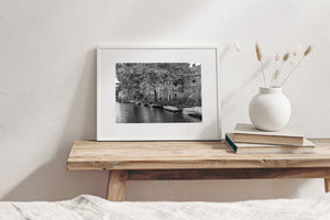 Monochrome canal photograph with open boat interiors, folded covers and historic buildings partly hidden by leaves.