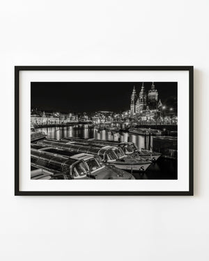 Glass-roofed canal boats and the illuminated Basilica of Saint Nicholas fill an Amsterdam night scene in black and white.
