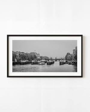 Boats, bridge structures and waterfront buildings span an Amsterdam canal in a black and white panorama.