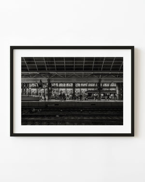 Travellers wait beneath the roof grid at Amsterdam Central Station across dark railway tracks in black and white.