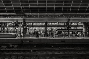 Small commuter figures gather on a bright platform beneath repeating station roof panels and beams.