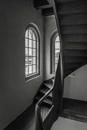 Light from tall arched windows meeting a dark timber staircase in an Amsterdam building.
