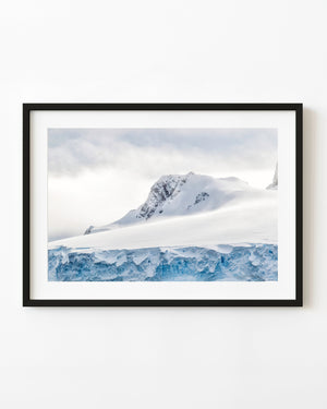 Blue Antarctic ice cliff beneath a snowy mountain peak and pale clouded sky.