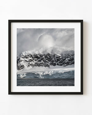 Snow-covered Antarctic cliffs beneath a large billowing cloud above dark coastal water.