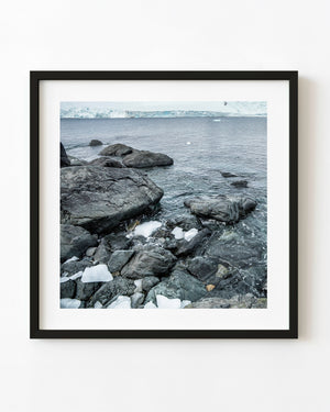 Dark coastal rocks and white ice fragments beside the Southern Ocean with an Antarctic glacier in the distance.